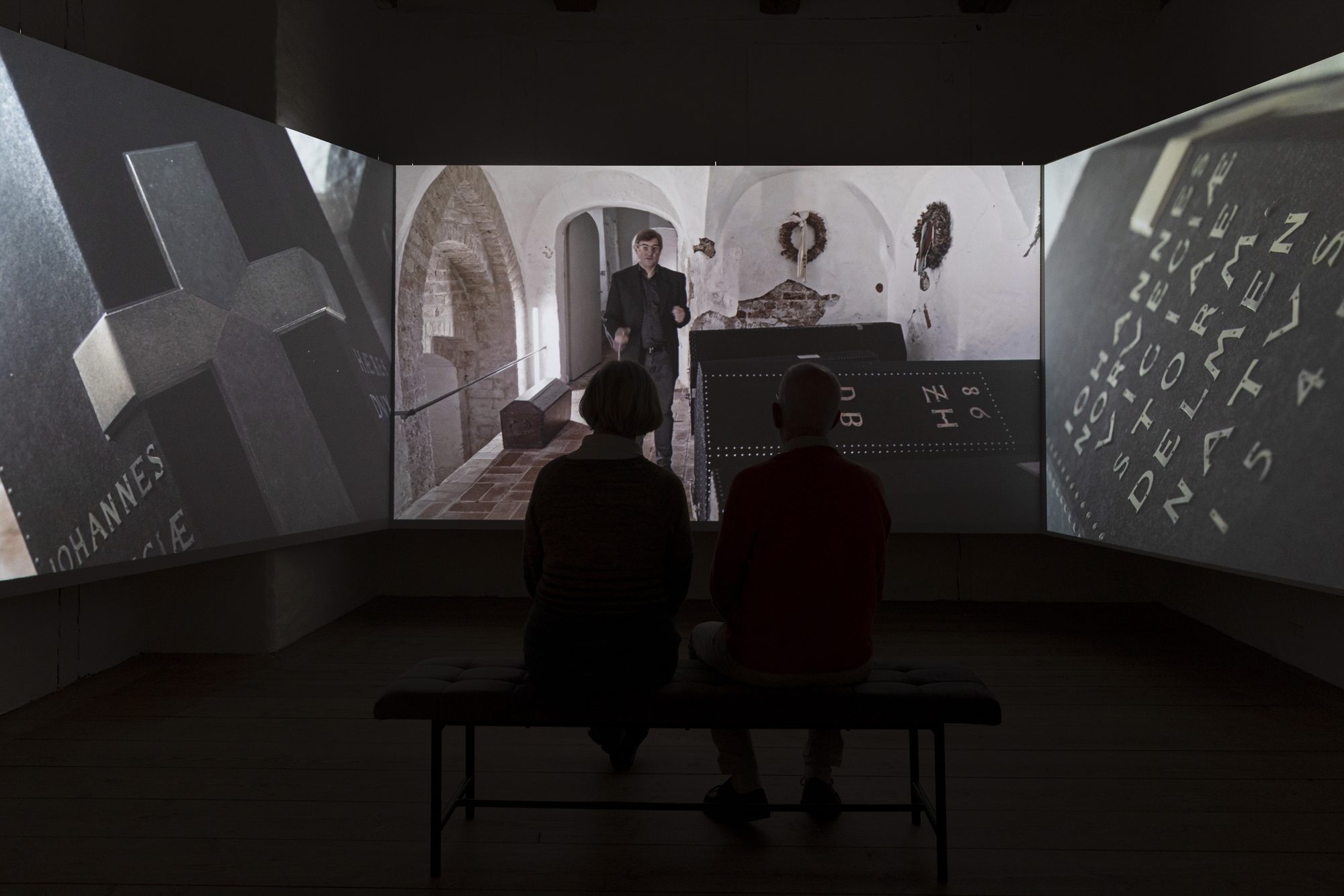 Makabre Mesterværker, visitors on a bench in front of the projected burial chapel and cross
