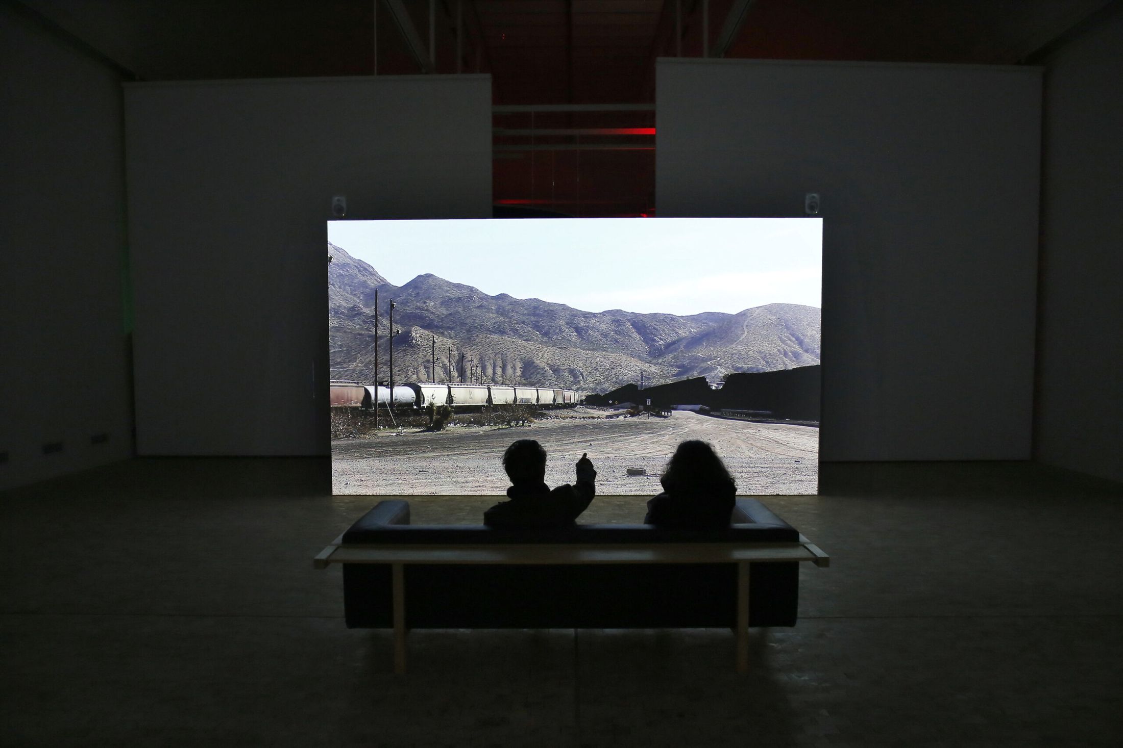 Membrane, two visitors on a bench in front of the video of the border wall